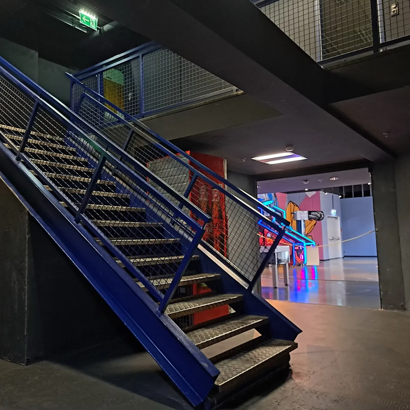 Eine Innentreppe mit blauen Geländern und Metallstufen, die zu einer oberen Ebene mit einem Neonschild und einer farbenfrohen Wandmalerei führt.