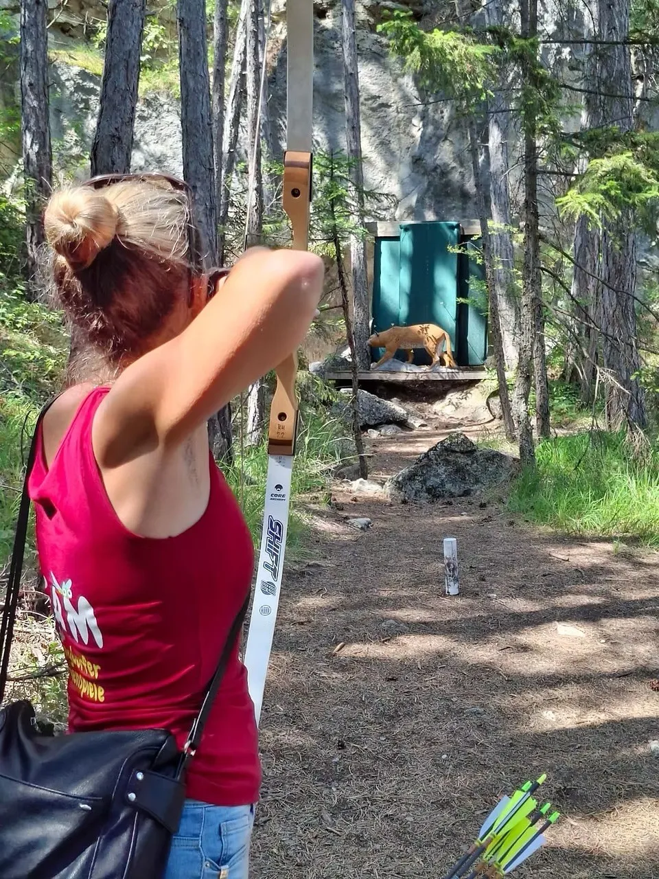 Eine Frau in einem rosa Tanktop übt Bogenschießen im Wald. Ein Puma befindet sich in der Nähe auf einer Plattform.
