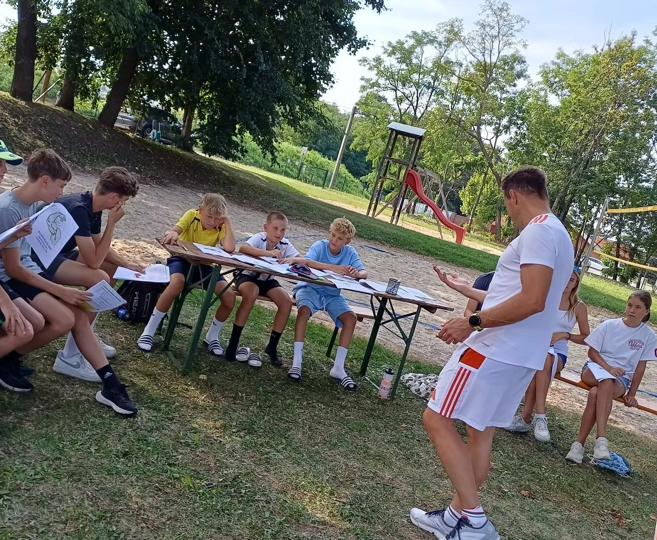 Eine Gruppe junger Jungen sitzt an einem Tisch im Park und schreibt auf Papiere. Ein Mann in einem weißen Shirt steht und spricht mit ihnen.