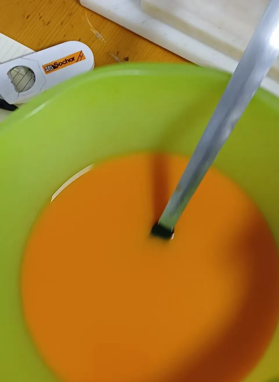 Ein grüner Behälter mit orangefarbener Flüssigkeit, darin ein Metalllöffel. Ein weißes Objekt mit dem Wort 'Sochor' steht neben dem Behälter.
