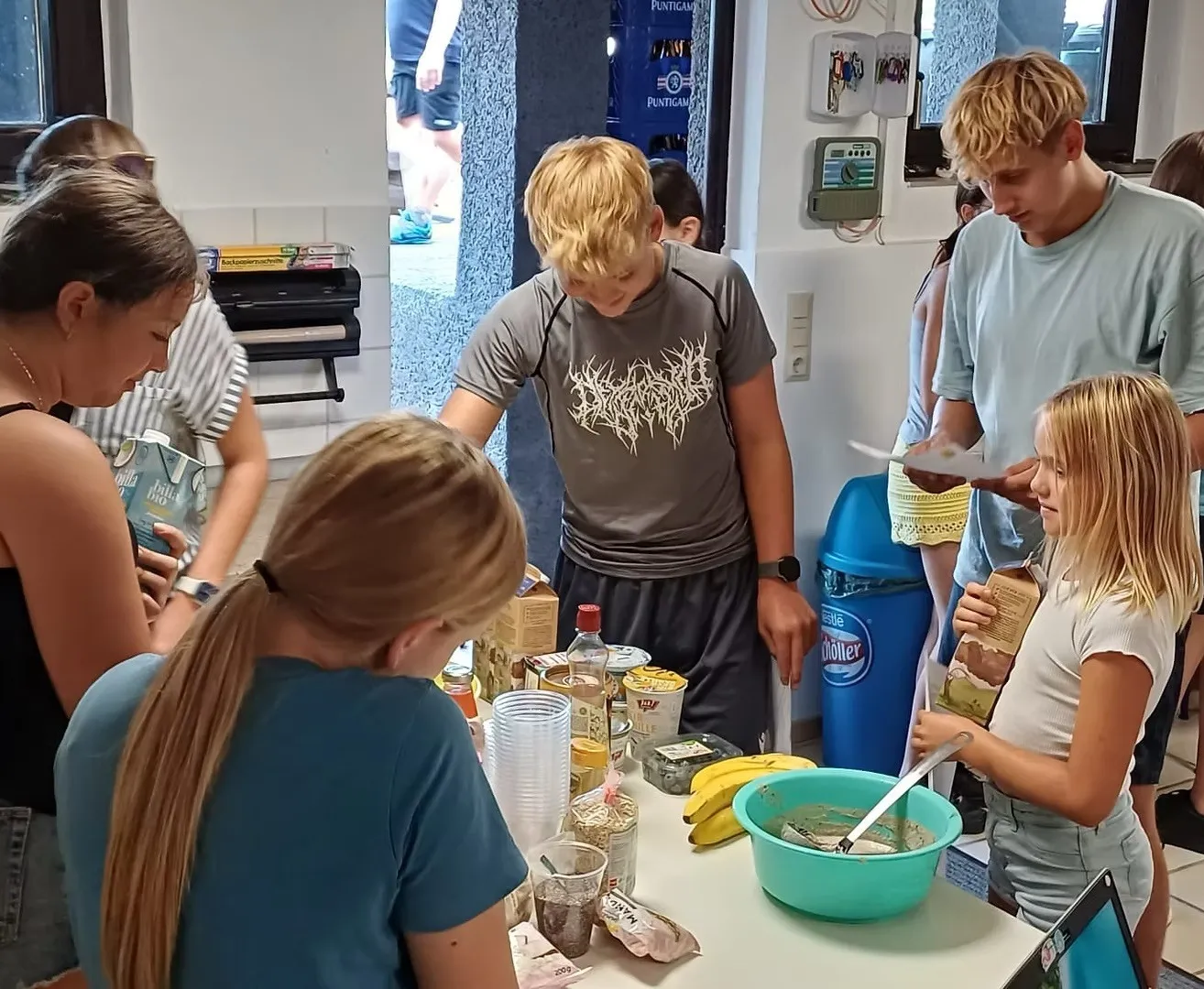 Eine Gruppe von Kindern versammelt sich um einen Tisch mit verschiedenen Lebensmitteln und einer Schüssel, möglicherweise bereiten sie gemeinsam eine Mahlzeit zu.