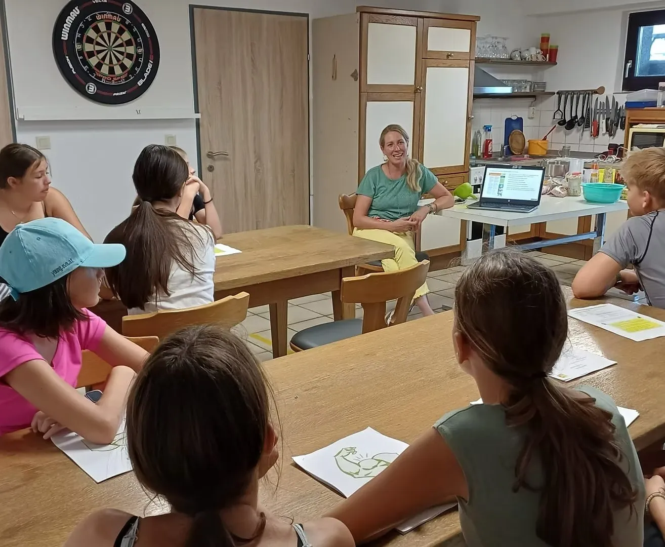 Eine Frau sitzt an einem Laptop in einem Klassenzimmer und unterrichtet eine Gruppe von Mädchen. Die Mädchen sitzen an Tischen, einige mit Papieren und Stiften. Ein Dartboard ist an der Wand.