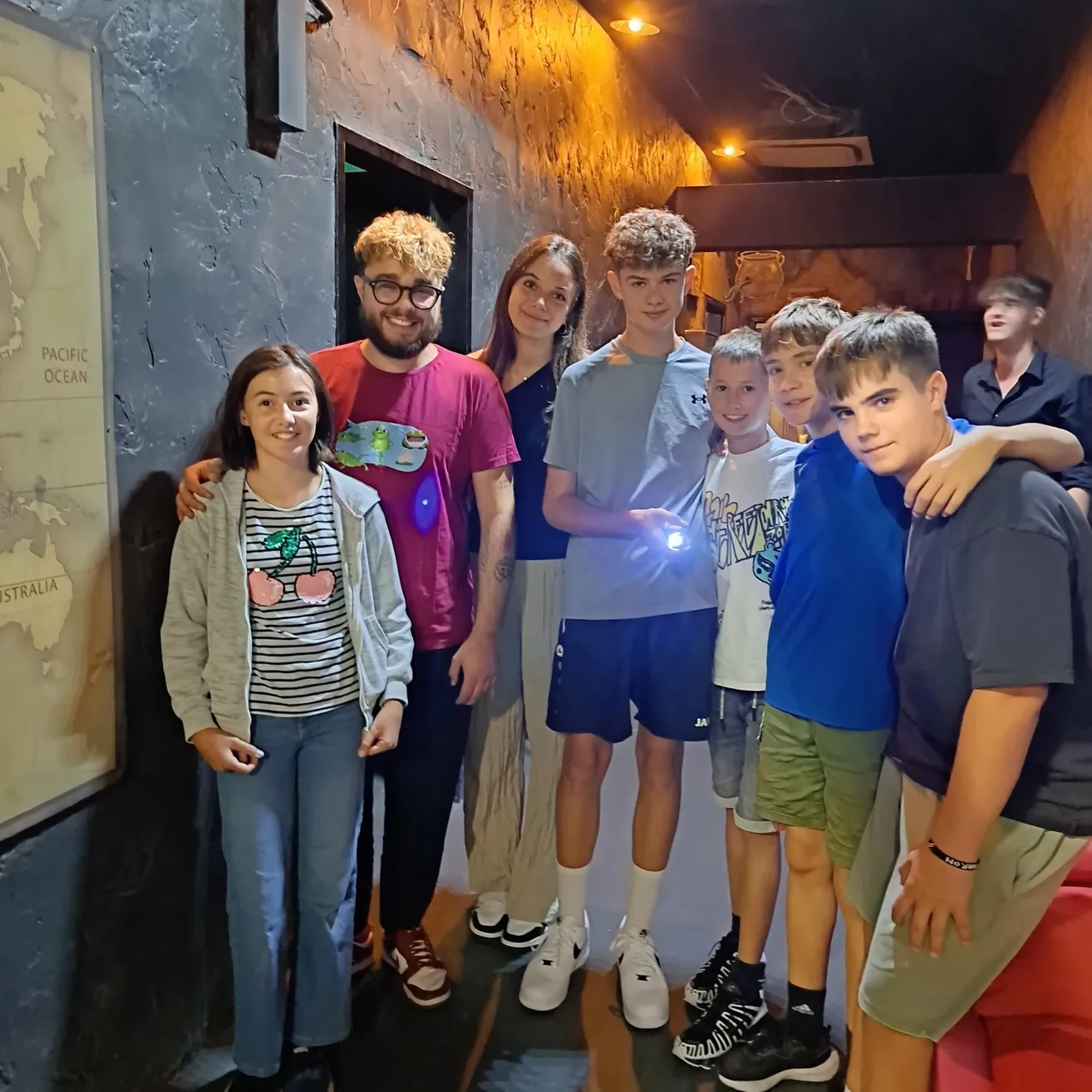 Eine Gruppe junger Menschen steht in einem schwach beleuchteten Raum und lächelt für ein Foto. Eine Karte des Pazifischen Ozeans ist an der Wand hinter ihnen sichtbar.