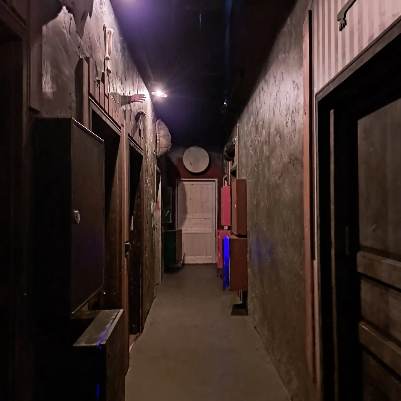 Ein schwach beleuchteter Korridor mit einer Hand, die aus der Wand ragt, beleuchtet von einer einzelnen Deckenlampe. Die Wände sind in einem abgenutzten Stil gestrichen, und es gibt verschiedene Türen und Schränke entlang der Seiten.