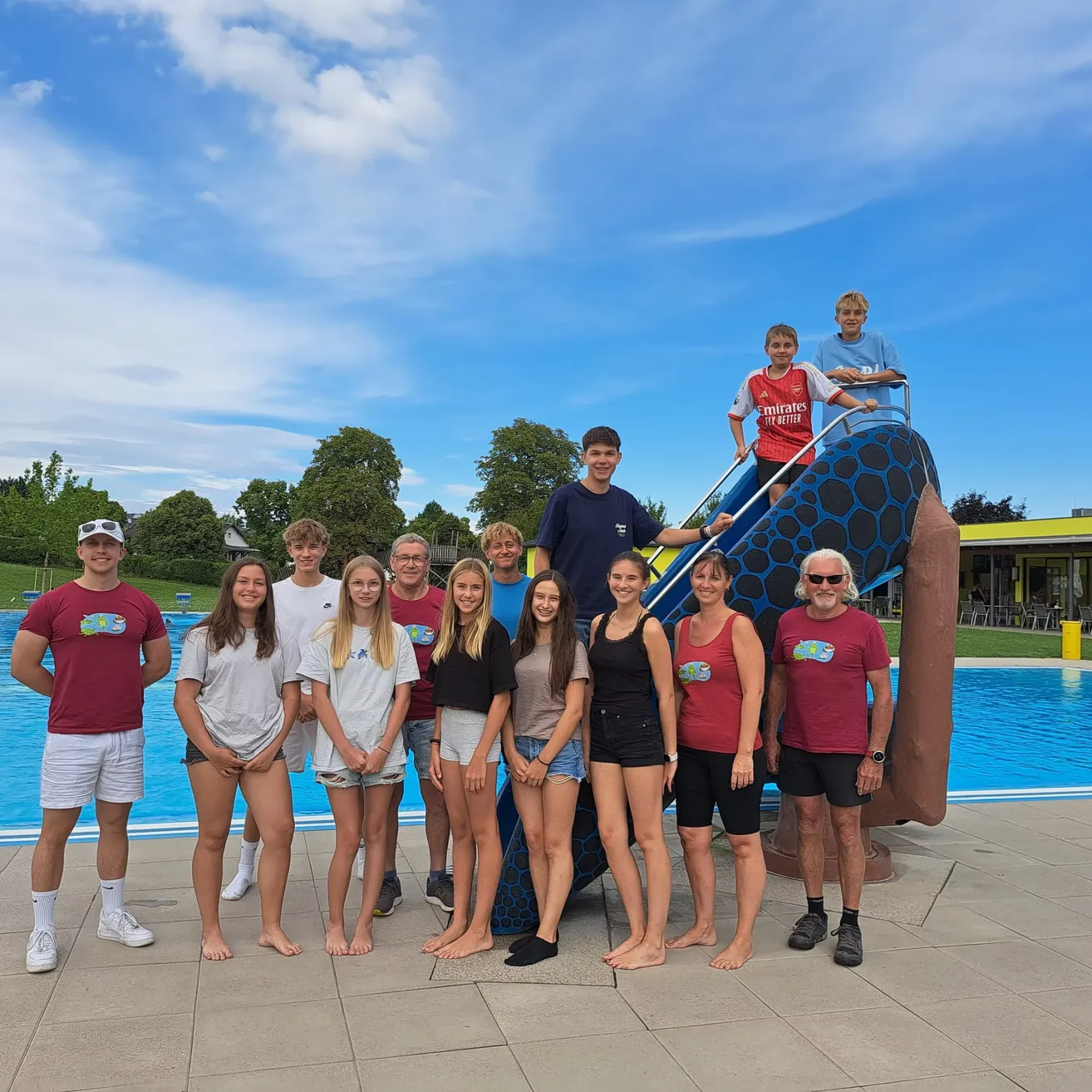 Eine Gruppe junger Menschen und Erwachsener steht vor einem Pool, wobei zwei Jungen auf einer Rutsche im Hintergrund zu sehen sind.