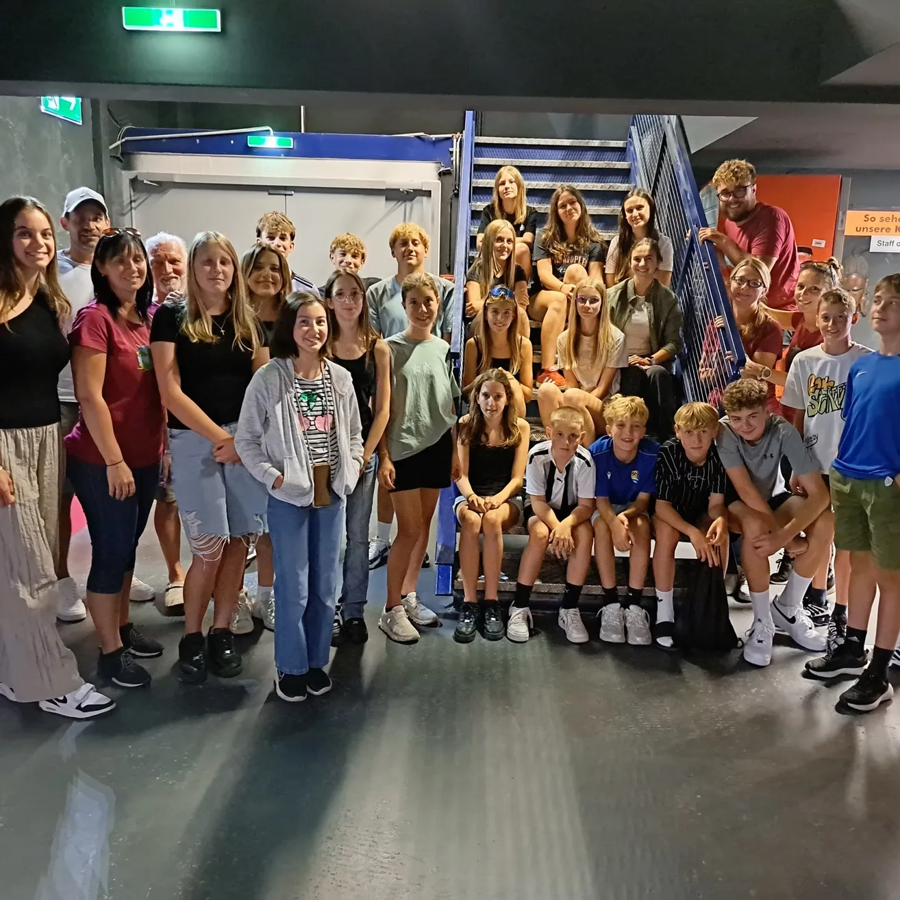 Eine Gruppe von Menschen, einschließlich Erwachsener und Kindern, posiert für ein Foto in einem Raum mit Treppen und einer Wand mit einem Schild.