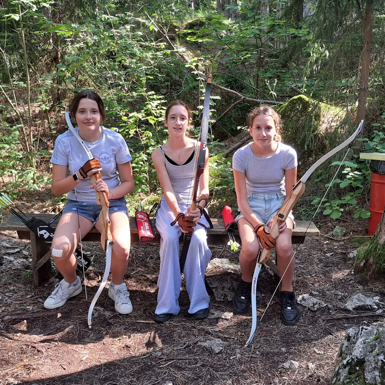 Drei junge Frauen sitzen auf einer Bank im Wald, jede hält einen Bogen und Pfeil. Sie lächeln und posieren für ein Foto.