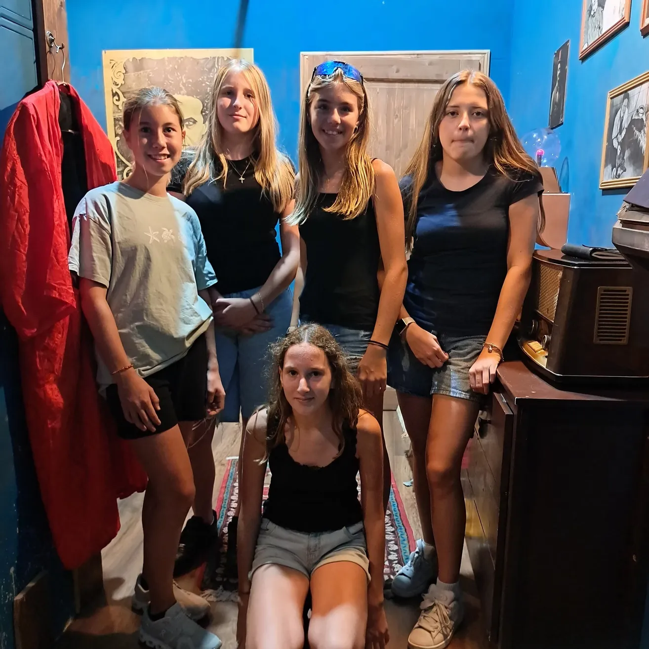 Fünf junge Mädchen stehen in einem blauen Raum zusammen und lächeln für ein Foto. Ein Mädchen kniet, und ein anderes trägt eine Sonnenbrille auf dem Kopf.