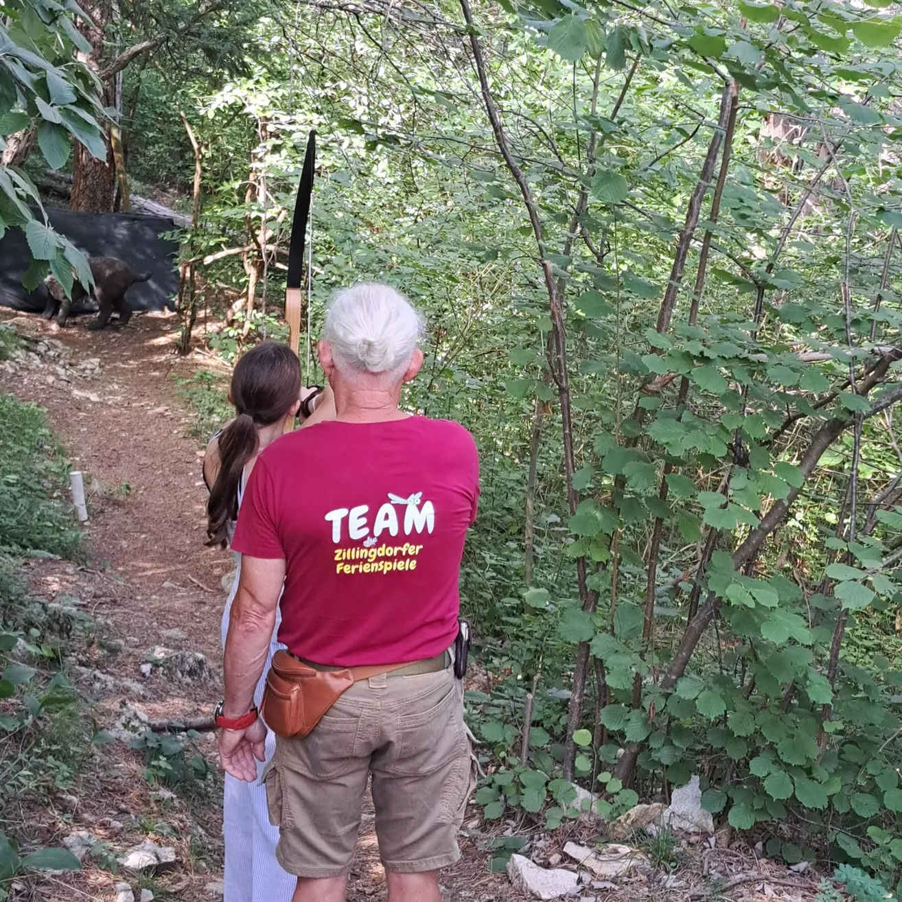 Ein älterer Mann mit einem roten T-Shirt und einem Bogen geht mit einer Frau durch einen Waldweg.