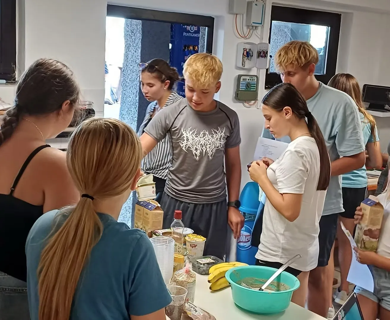 Eine Gruppe junger Menschen versammelt sich in einer Küche um einen Tisch und bereitet Essen zu. Ein Junge in einem grauen T-Shirt greift nach etwas auf dem Tisch. Ein Mädchen in einem weißen T-Shirt hält einen Löffel.