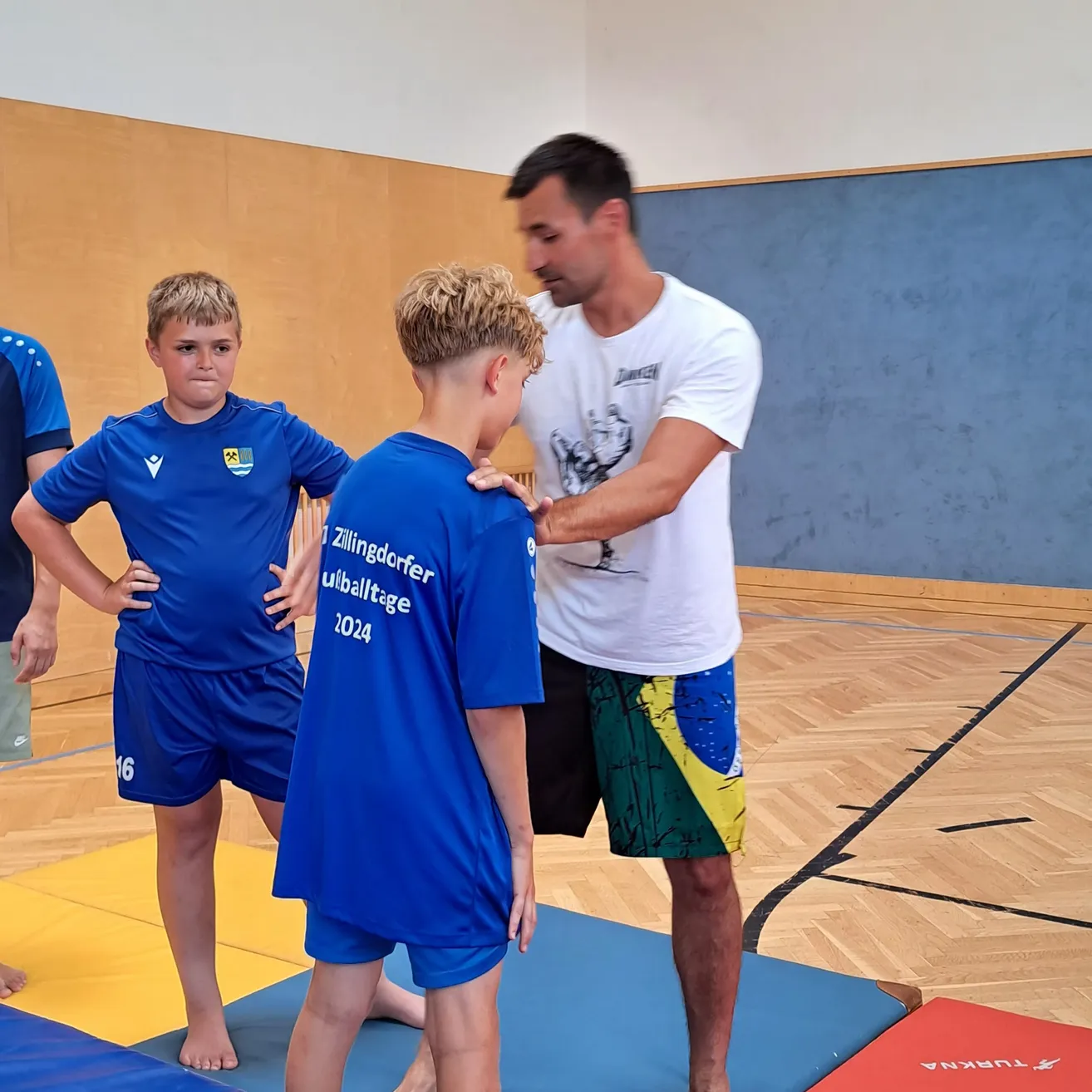 Ein Mann in einem weißen T-Shirt steht zwischen zwei Jungen in blauen Uniformen, möglicherweise in einer Turnhalle. Die Jungen sind barfuß und stehen auf blauen Matten.