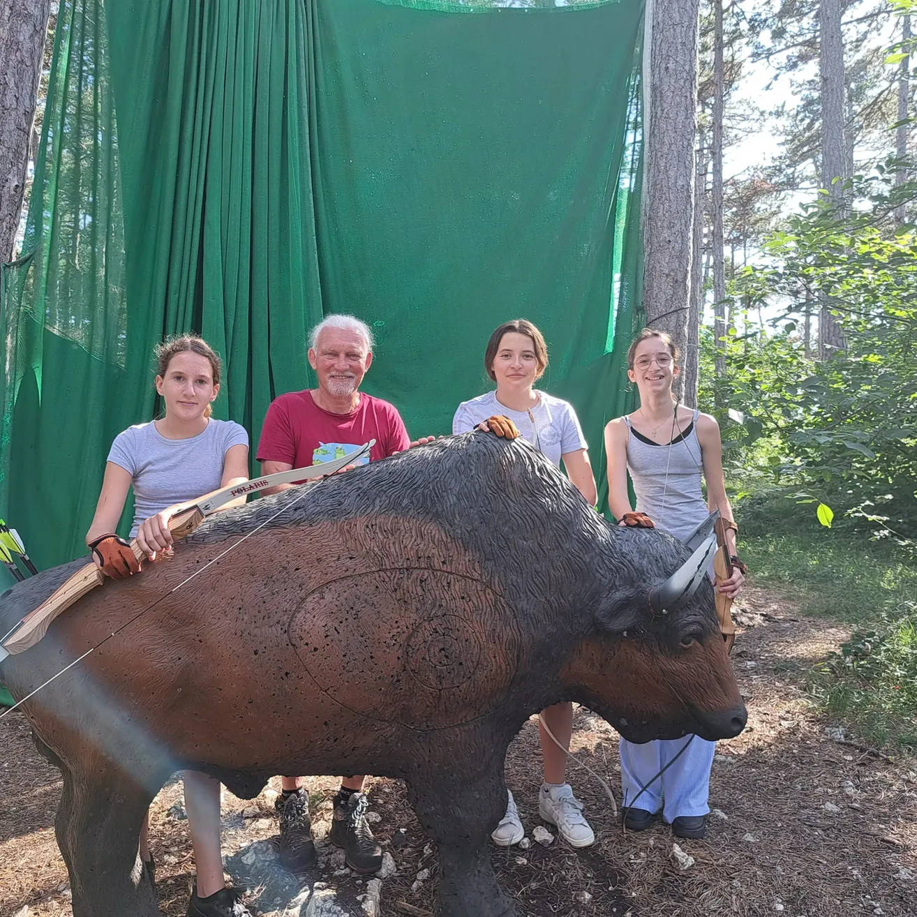 Eine Gruppe von vier Personen, zwei Frauen und ein älterer Mann, posiert mit einer großen hölzernen Bisonstatue im Wald. Sie halten Bögen und Pfeile.