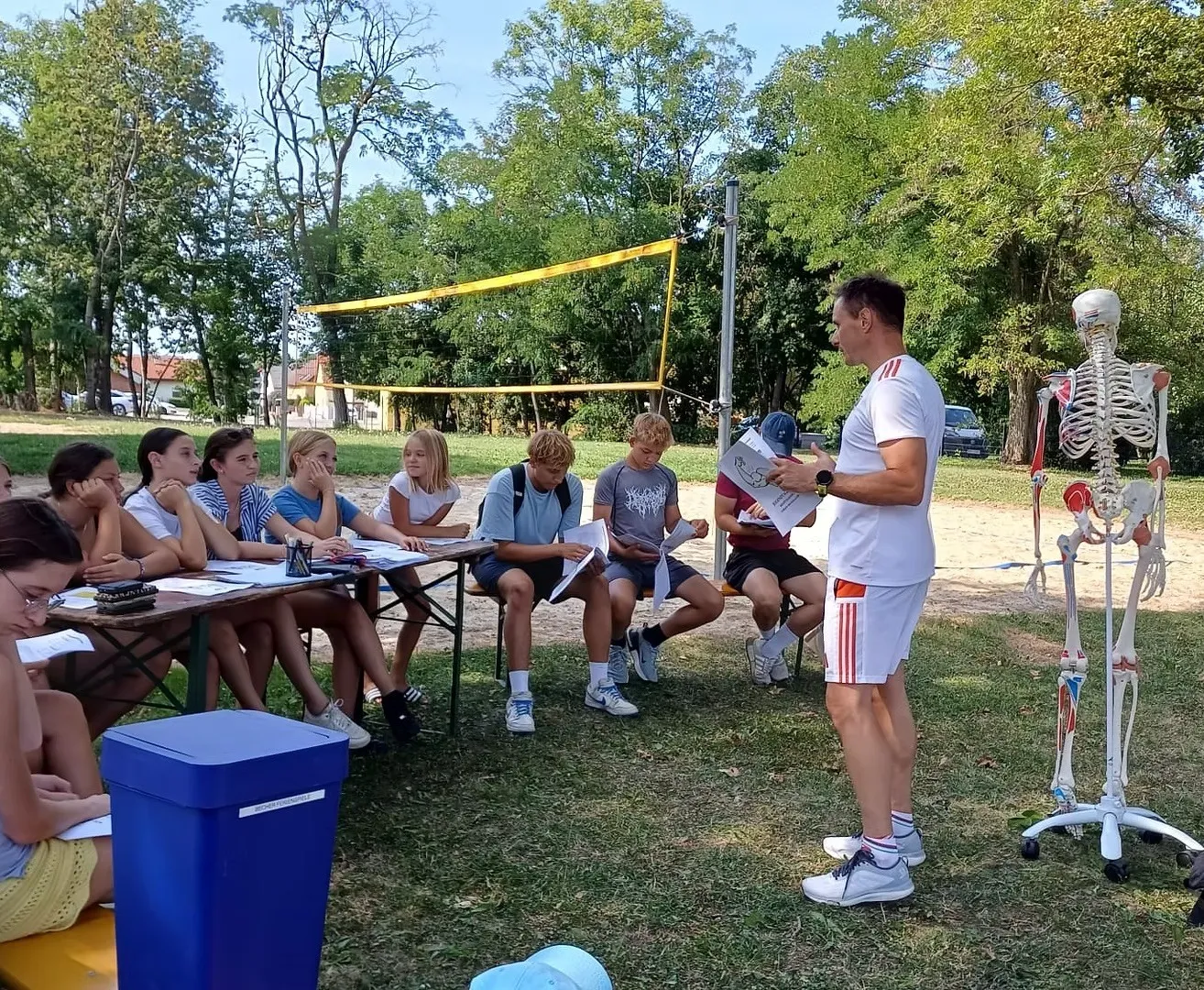 Eine Gruppe von Schülern sitzt auf Bänken in einem Grasbereich und hört einem Mann zu, der vor ihnen steht. Der Mann hält ein Stück Papier in der Hand und scheint etwas zu erklären. Ein Volleyballnetz ist hinter ihnen aufgestellt, und ein Skelettmodell ist auf der rechten Seite platziert.