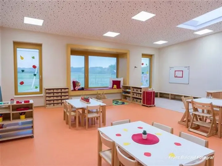 Ein Klassenzimmer mit weißen Tischen und Stühlen, einer Fensterbank und einer Tafel. Der Raum hat ein helles, luftiges Gefühl mit orangefarbenem Boden und weißen Wänden.