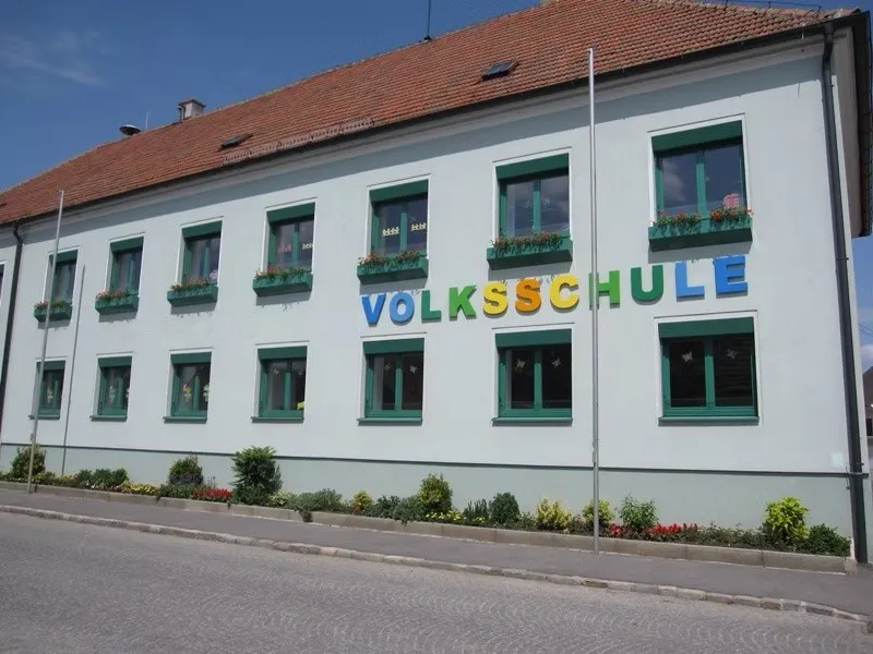 Ein weißes Gebäude mit rotem Dach und grünen Fensterrahmen. Es hat das Wort 'Volksschule' in Blau an der Wand geschrieben. Es gibt Blumenkästen an den Fenstern.