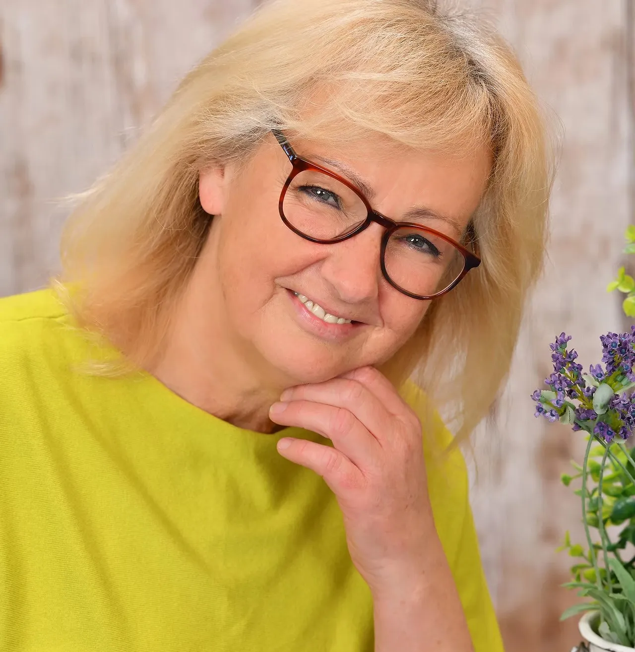 Eine Frau mit blonden Haaren, die eine Brille und ein gelbes T-Shirt trägt, lächelt und posiert für ein Foto. Sie hat ihre Hand am Kinn.