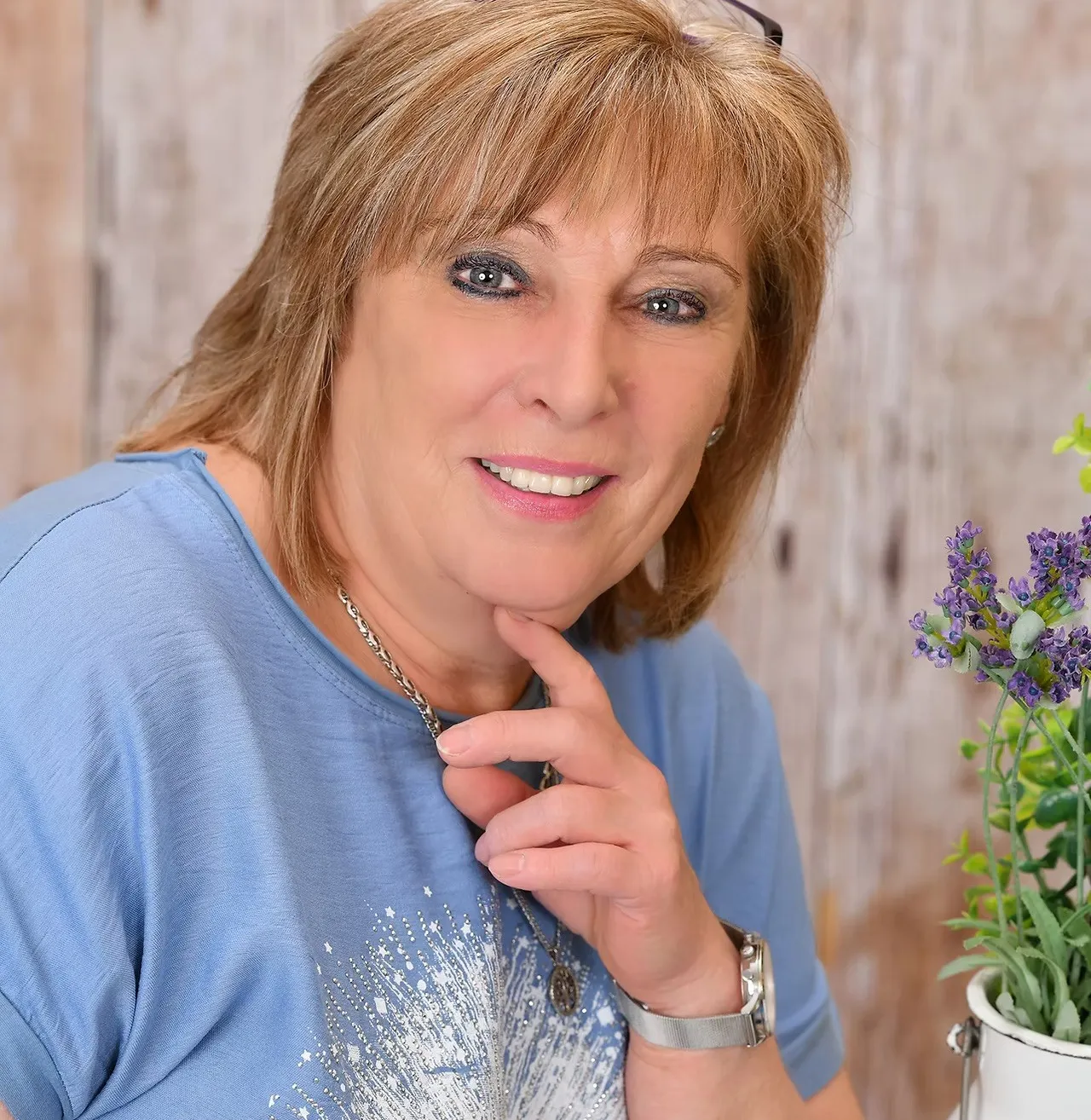 Eine Frau mit kurzen blonden Haaren lächelt und posiert für ein Foto. Sie trägt ein blaues Shirt und eine Halskette mit Anhänger. Sie berührt ihr Kinn mit der rechten Hand. Hinter ihr befindet sich eine Holzwand und ein Topf mit Lavendelblumen.