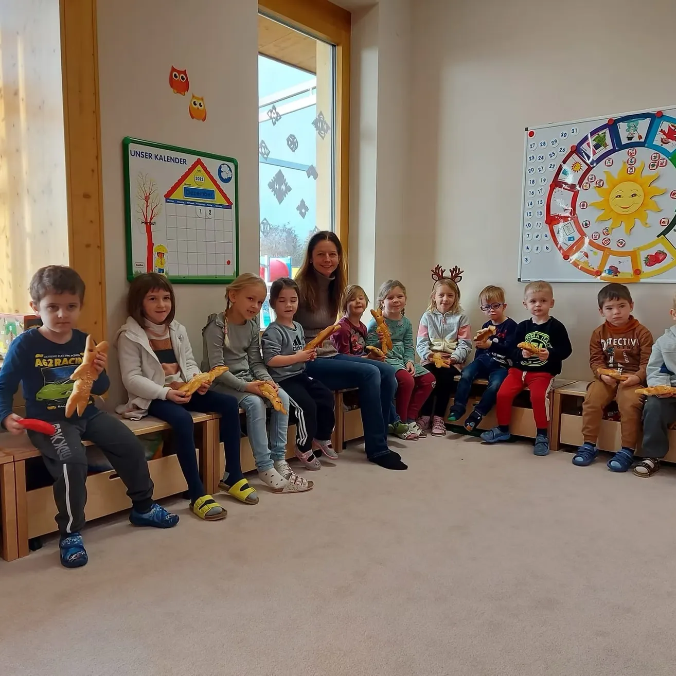 Eine Gruppe von Kindern und einer erwachsenen Frau sitzen auf Bänken in einem Raum, halten Brot und lächeln. Ein Kalender und ein kreisförmiges Diagramm hängen an der Wand.