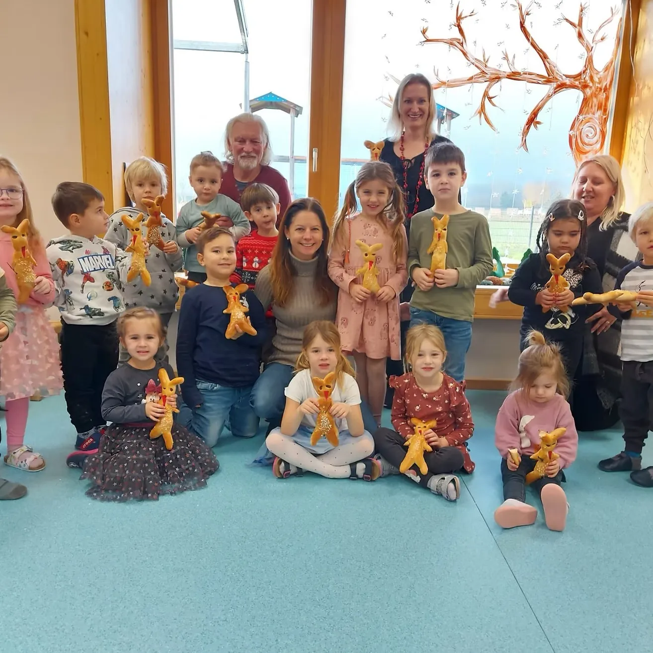 Eine Gruppe von Kindern und Erwachsenen posiert für ein Foto und hält Plüsch-Känguru-Spielzeug. Sie sind in einem Raum mit großen Fenstern und einer Baummalerei.