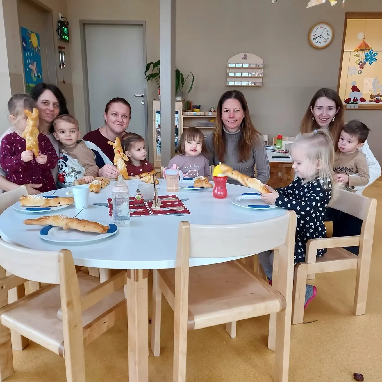 Eine Gruppe von Erwachsenen und Kindern versammelt sich um einen Tisch, isst Brot und lächelt. Der Tisch ist mit Tellern, Tassen und einer Flasche gedeckt. Der Raum hat eine warme, gemütliche Atmosphäre.