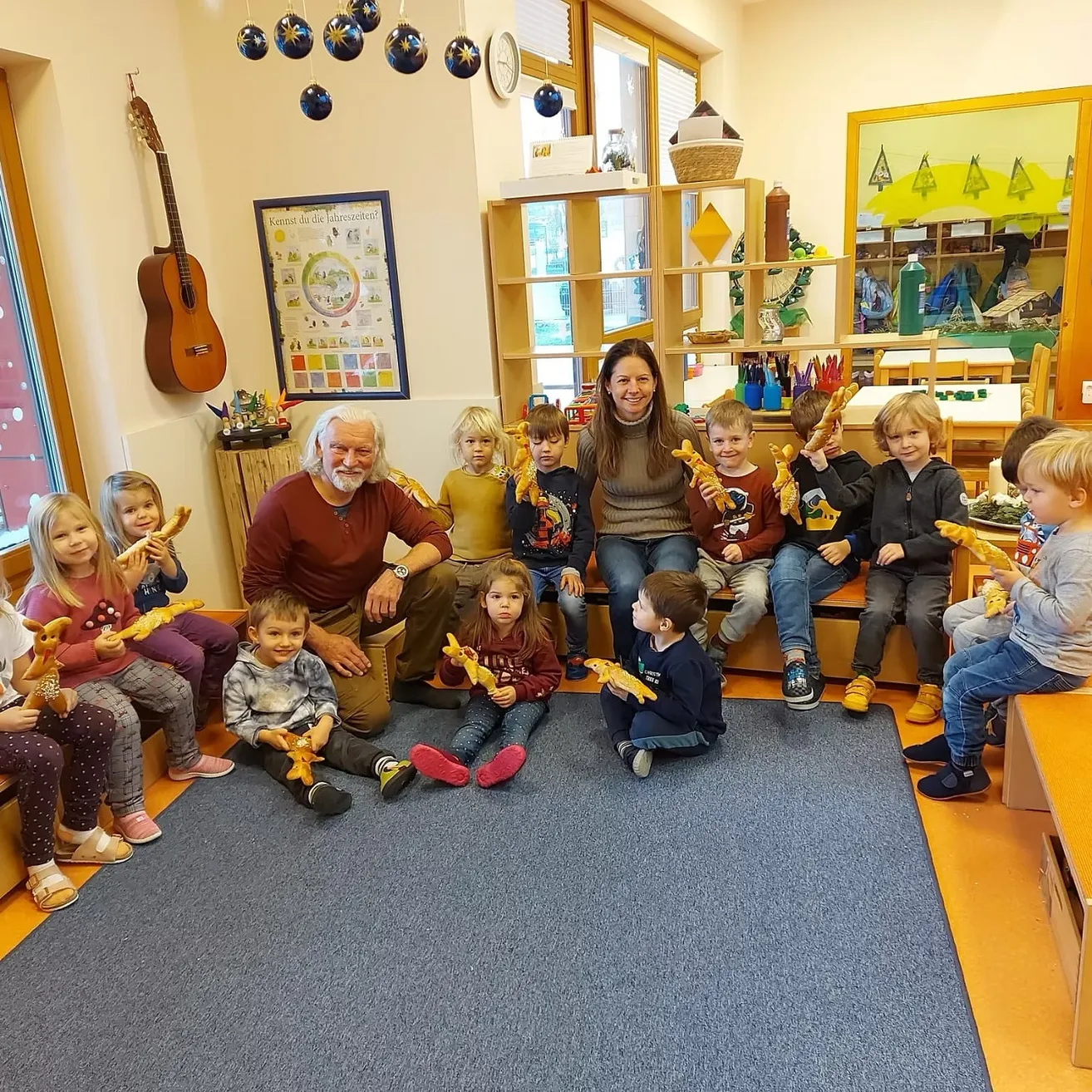 Eine Gruppe von Kindern und zwei Erwachsenen sitzen auf einer Bank in einem Raum und halten Spielzeug. Der Raum hat einen Holzboden, eine Wand mit einer Uhr und ein Regal mit Spielzeug.
