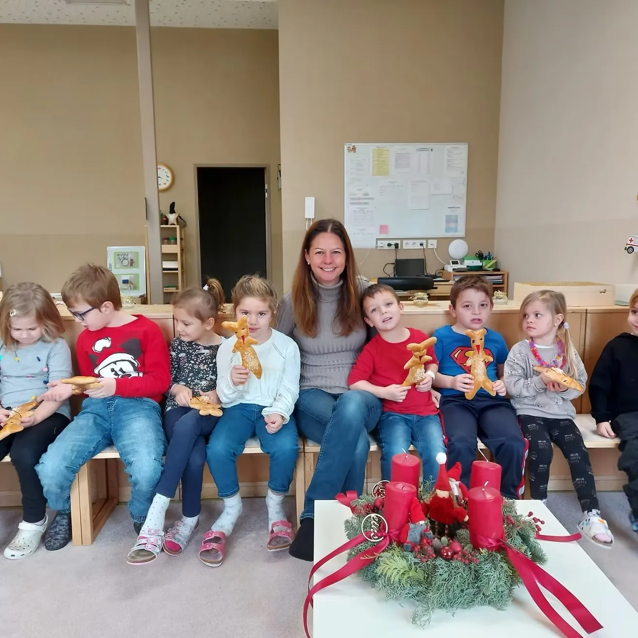 Eine Frau und mehrere Kinder sitzen auf einer Bank und halten Lebkuchen. Sie sind in einem Raum mit einem Weihnachtskranz und Kerzen auf einem Tisch vor ihnen.