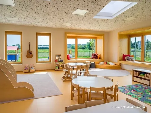 Ein heller und geräumiger Spielraum mit Holzmöbeln, großen Fenstern und Blick auf ein grünes Feld. Der Raum verfügt über eine Gitarre an der Wand, einen Teppich und einen Tisch mit Stühlen.