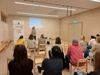 Eine Frau steht vorne in einem Raum und spricht zu einem sitzenden Publikum. Der Raum hat Holzstühle, eine Projektionsleinwand und ein Schild an der Wand.