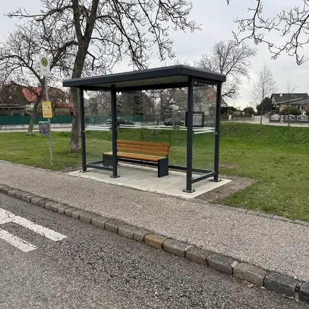Eine leere Bushaltestelle mit einer Holzbank befindet sich auf einem gepflasterten Weg in der Nähe eines Rasens.