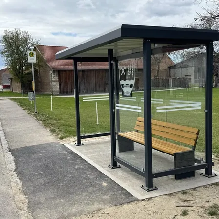 Eine moderne Bushaltestelle mit Glasverkleidung, einer Holzbank und einem Logo auf der Scheibe. Sie befindet sich auf einem Betonpodest in der Nähe eines Rasenfeldes.