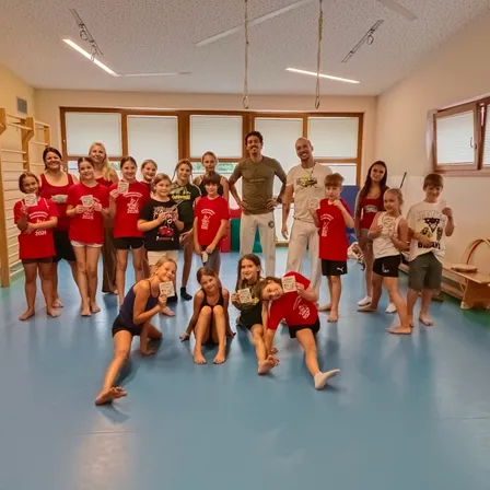 Eine Gruppe von Kindern und Erwachsenen in roten T-Shirts posiert für ein Foto in einem Fitnessstudio. Sie halten Urkunden und lächeln. Zwei Männer in grünen und weißen T-Shirts stehen hinter ihnen.