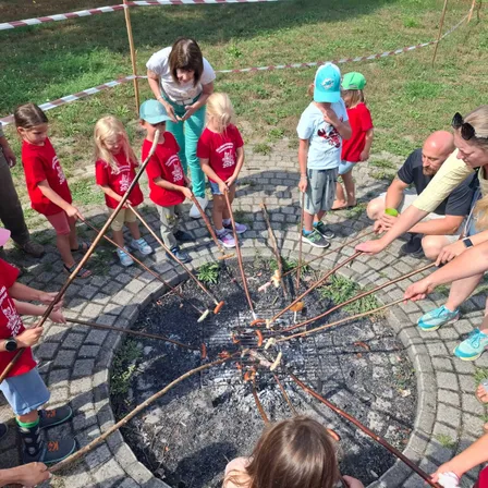 Eine Gruppe von Kindern und Erwachsenen versammelt sich um ein Feuer, um Würste an Stöcken zu rösten. Sie tragen rote Hemden und Mützen.