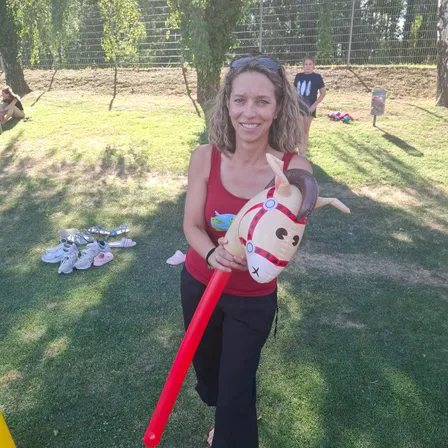 Eine Frau in einem roten Tanktop hält ein rotes aufblasbares Pferd auf einem Rasen. Andere Personen und Gegenstände sind im Hintergrund zu sehen.