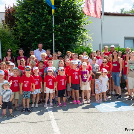 Eine Gruppe von Kindern und Erwachsenen in roten T-Shirts mit dem Jahr 2026, die vor einem Gebäude stehen.