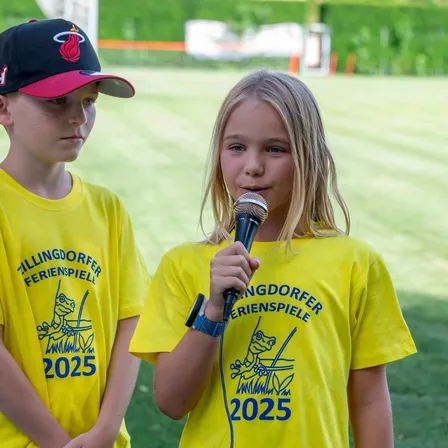 Zwei Kinder in gelben T-Shirts mit dem Jahr 2025 darauf. Eines hält ein Mikrofon und das andere schaut zu. Sie stehen auf einem Rasenfeld.