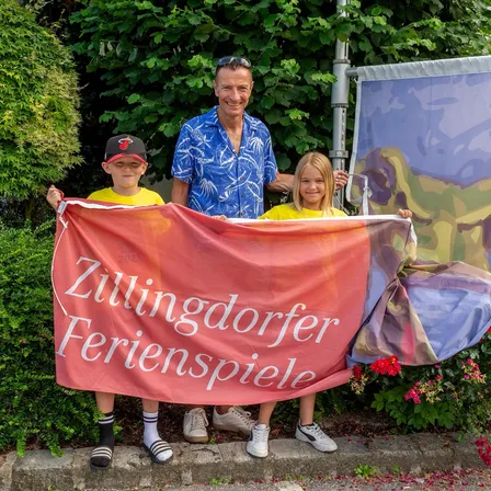 Ein Mann und zwei Kinder stehen vor einem Banner mit der Aufschrift 'Zulingdorfer Ferienspiele'. Der Mann trägt ein blaues Hemd und eine Sonnenbrille auf dem Kopf. Die Kinder tragen Turnschuhe und halten das Banner. Dahinter befindet sich ein farbenfrohes Banner und ein Busch mit grünen Blättern.