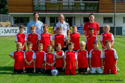 Eine Gruppe junger Fußballer in roten und weißen Trikots posiert mit ihren Trainern auf einem Spielfeld.