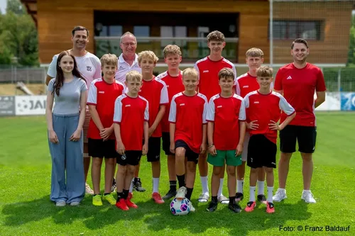 Eine Gruppe junger Fußballspieler in roten und weißen Trikots posiert für ein Foto auf einem Rasenfeld. Sie werden von zwei Erwachsenen begleitet, einer mit einer Brille und der andere mit einer Uhr. Das Foto ist mit 'Foto:' im unteren rechten Eck versehen.