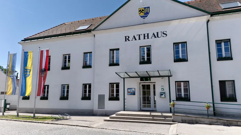 Abschnittsbild: Die Vorderseite eines weißen Gebäudes mit dem Wort Rathaus darauf. Es hat mehrere Fenster, einen Glasvorbau über dem Eingang und eine kleine Pflanze auf dem Geländer.