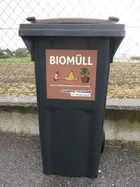 Ein schwarzer Mülleimer mit einem braunen Etikett, das 'BIOMUELL' liest. Das Etikett enthält Symbole eines Vogels, eines Wurms und einer Pflanze. Der Eimer steht auf einer Betonfläche mit einem Zaun im Hintergrund.