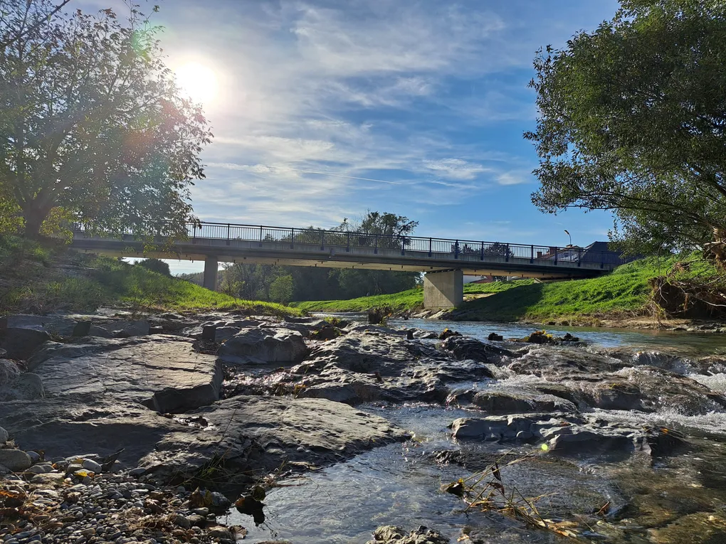 Abschnittsbild: Eine Brücke überspannt einen felsigen Fluss unter einem hellen, sonnigen Himmel. Bäume säumen die Ufer, und einige Leute gehen auf der Brücke.