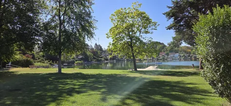 Eine ruhige Seelandschaft mit einem üppigen grünen Rasen, einem Baum und einem Steg, der sich in das ruhige Wasser erstreckt. In der Ferne säumen Häuser und Bäume den Uferrand.