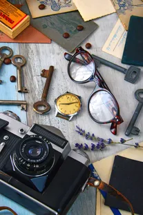 Eine Vintage-Kamera, Schlüssel, eine Uhr und eine Brille sind auf einer Holzoberfläche angeordnet. Die Kameralinse ist geöffnet, und die Uhr zeigt die Zeit 10:10 an.