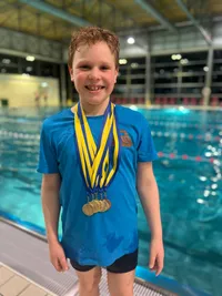 Ein junger Junge mit einem lächelnden Gesicht steht vor einem Schwimmbecken. Er trägt ein blaues T-Shirt mit einem Logo und mehrere goldene Medaillen um den Hals.