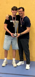 Ein junger Mann mit einer Medaille um den Hals hält eine Trophäe und steht neben einem lächelnden Mann.