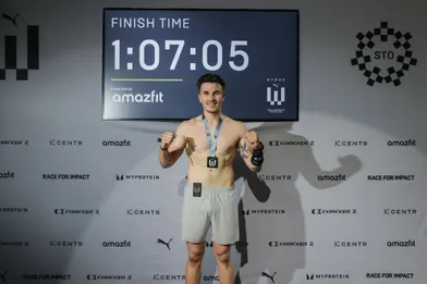 Ein Mann steht vor einer großen Anzeigetafel, die eine Endzeit von 1:07:05 anzeigt. Er trägt eine Medaille um den Hals und hat eine Tätowierung am Arm.