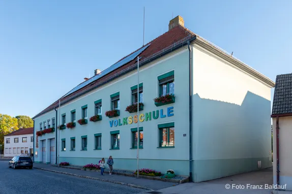 Ein weißes Gebäude mit grünem Rand und rotem Dach hat ein Schild, das Volksschule steht. Es gibt mehrere Fenster und eine Tür am Gebäude. Zwei Personen stehen auf dem Bürgersteig vor dem Gebäude.
