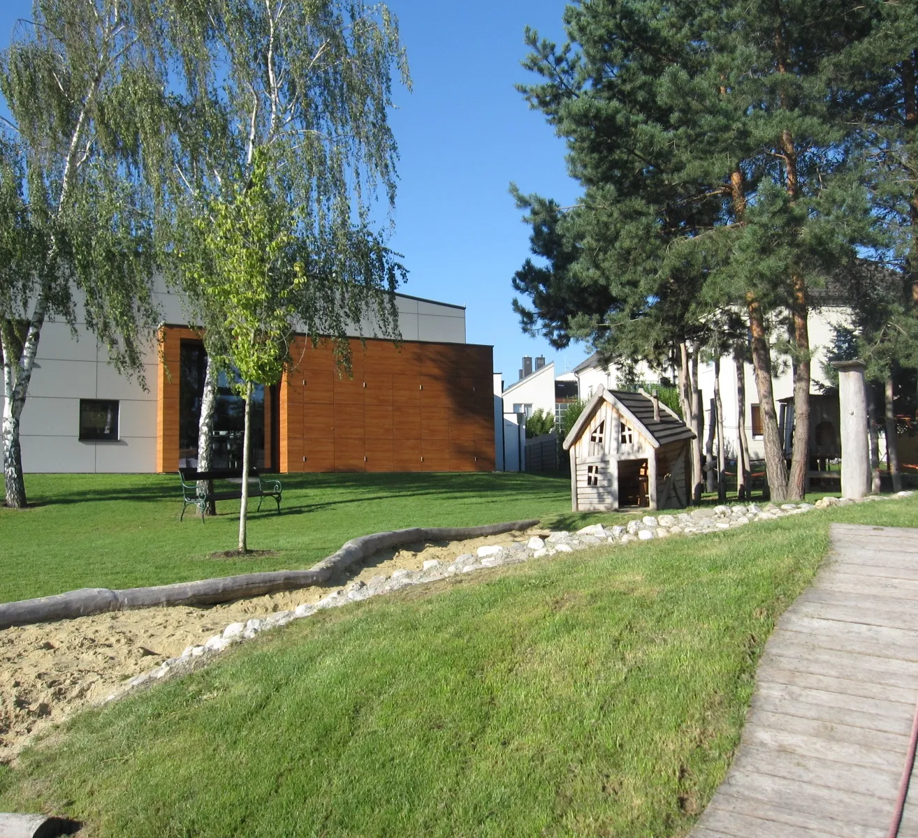 Ein Außenbereich mit einem Holzweg, der zu einem kleinen Holzhaus führt. Der Bereich ist von Bäumen und Gras umgeben.