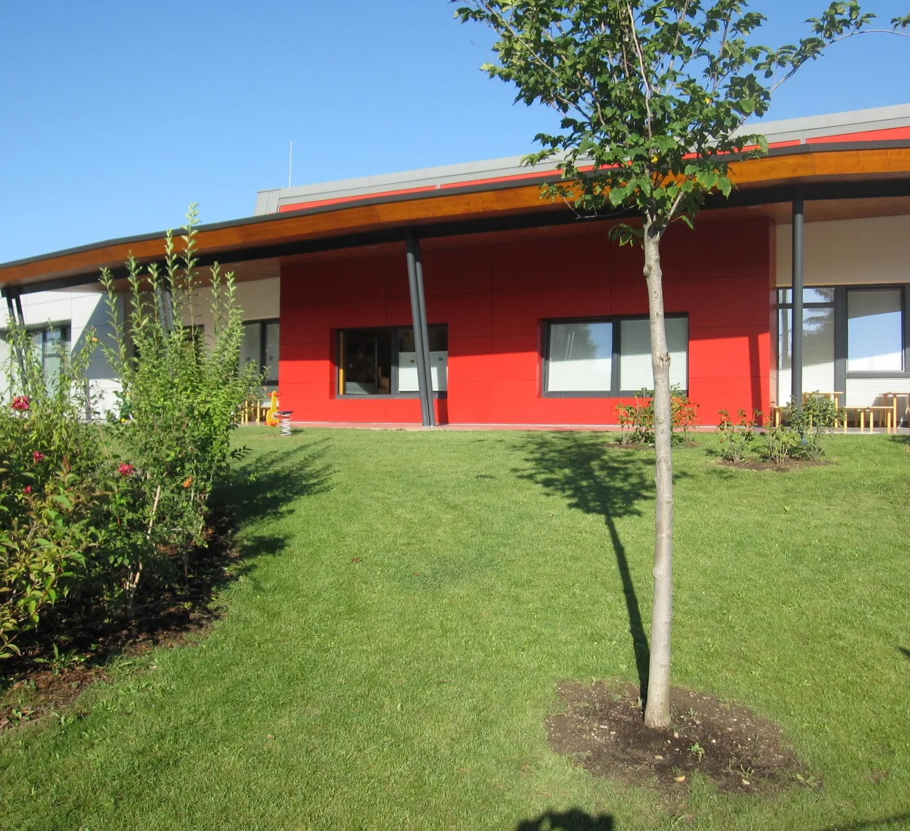 Ein modernes Gebäude mit roter Wand und weißen Rändern steht in einem gepflegten Garten. Das Gebäude hat ein geschwungenes Dach und große Fenster.