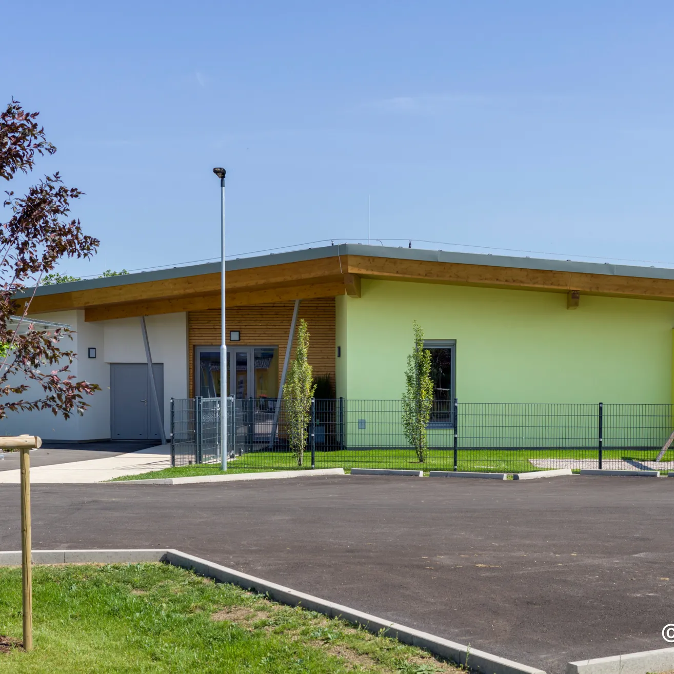 Ein modernes gelbes Gebäude mit einem grünen Dach, einem Parkplatz davor und einem Baum im Vordergrund.
